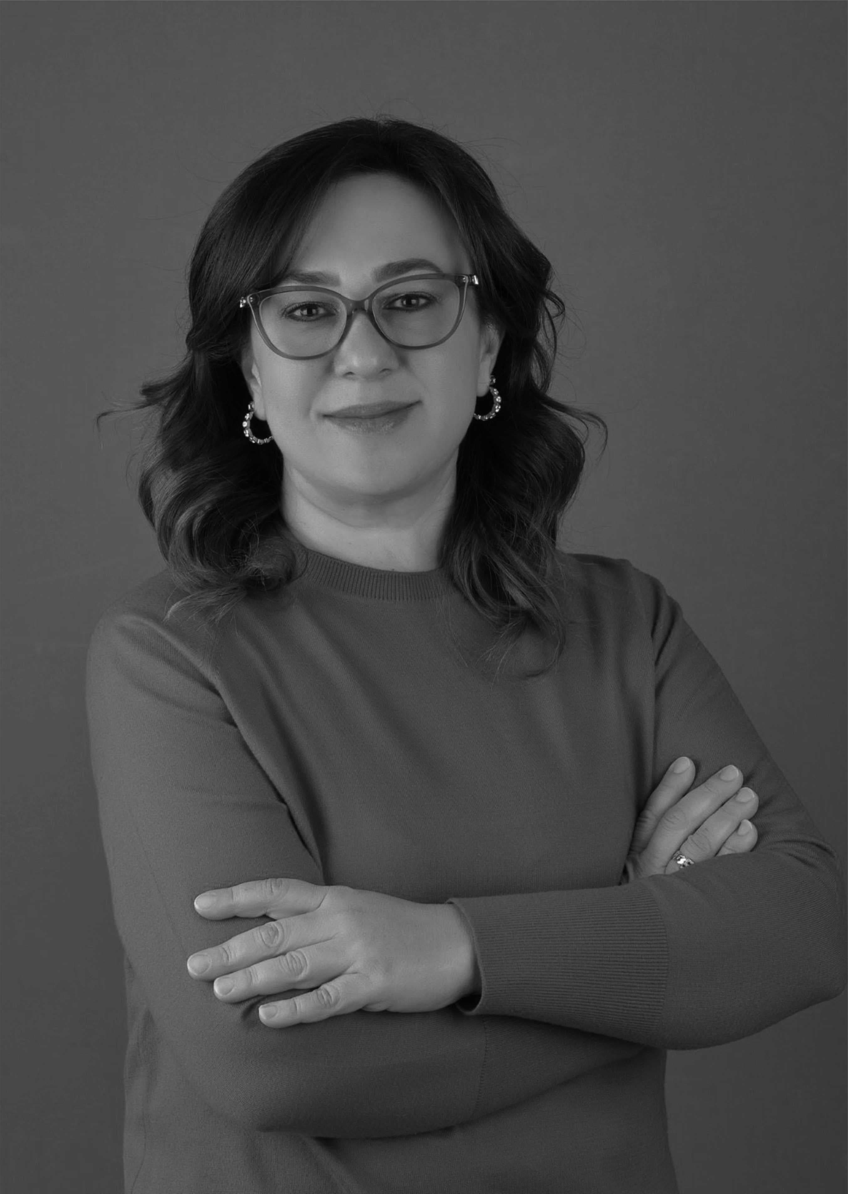 Smiling woman with glasses and wavy hair poses with arms crossed in monochrome.
